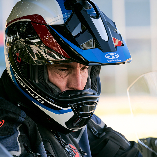 Close-up of a motorcyclist wearing a blue, white, and red full-face helmet with a visor. The helmet appears to be a Kabuto brand. The rider is also wearing dark motorcycle gear.